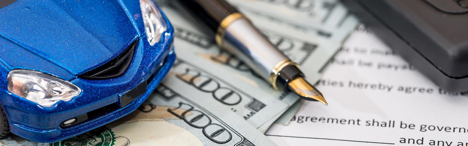 Close-up of a New Car Financing agreement with calculator, pen, hundred dollar bills and the front end of a bright blue toy vehicle on top of it.